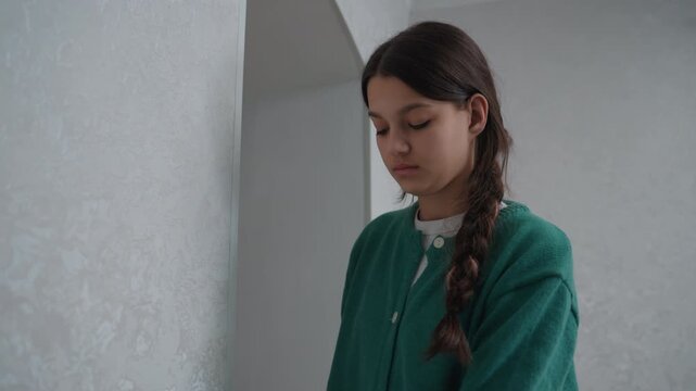 Young woman in empty apartment standing, wearing green cardigan and braided hair, quiet expression, soft daylight, newcomer reflecting after moving, minimalist plaster walls and muted tones,