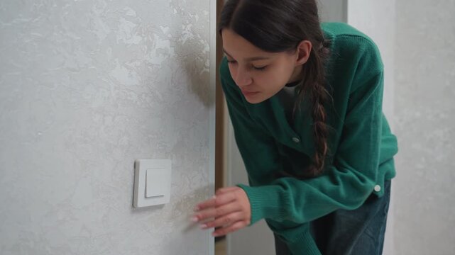 Young woman testing apartment light switch, careful hands on switch plate, braided hair and green cardigan, peeking from doorway to inspect wiring and power, practical movein check, textured plaster