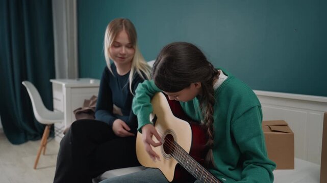 Two white roommates in new apartment sharing warm moment as one tunes acoustic guitar on couch amid cardboard boxes and teal accent wall, daylight, casual clothes, college students celebrating moving
