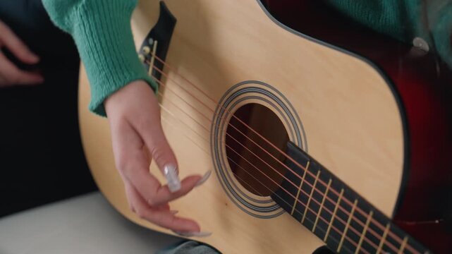 Closeup white hands strumming acoustic guitar with green sweater and pick, precise plucking across steel strings, wood rosette detail, soft natural light, focused practice session in new apartment,