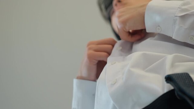 Anxious man fastening white shirt with trembling hands, lowangle closeup shows collar and cuff adjustment, shallow depth, stiff posture, preparing as job candidate outside office lobby, breathy pause
