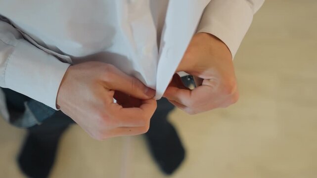 Close up caucasian man buttoning shirt groom overhead shot showing hands, white dress shirt, suspenders, wooden floor, gentle natural light, slow deliberate motion, nervous anticipation and ritual