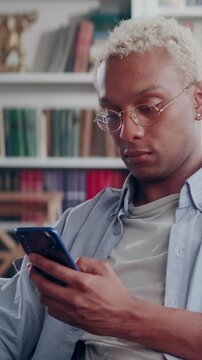 A blonde African American man with glasses looks intently and seriously at his smartphone screen while scrolling through updates or messaging in a room.