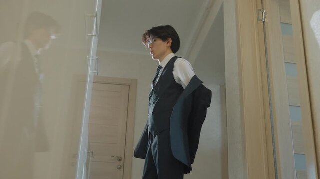 Young man putting on suit in hallway preparing for interview adjusting tie, checking mirror reflection, holding jacket and smartphone, tailored vest and cufflinks visible, morning sunlight