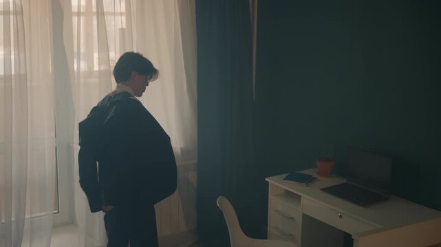 Young man contemplating in quiet room. Gentle morning light reveals thoughtful young man. Young man stands by window with jacket while reflecting on choices over soft dawn illumination