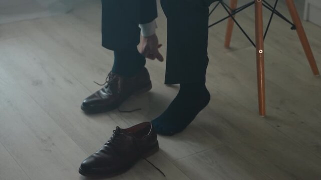 Man sitting putting on dress shoes, careful hands tying laces of polished brown leather beside chair on laminate floor, suit trousers and visible cuff, focused morning routine for commute or meeting,