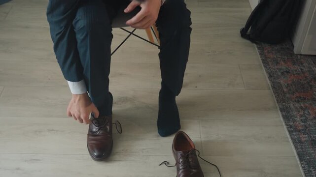 White businessman tying dress shoes on hardwood floor, bending from chair to lace polished brown brogues frames depict morning routine and focused executive preparing for interview, young lawyer