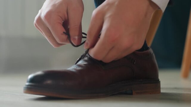Intimate closeup brown shoe being tied, calm morning preparation with hands fastening leather oxford on wooden floor near chair polished detail, classic brogue texture, slow cinematic motion conveying
