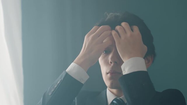 Asian businessman adjusts hair in suit, moody window light and soft cinematic haze corporate executive rehearses boardroom presentation, tailors tie, checks reflection with composed focus and quiet