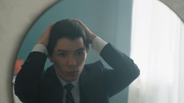 Asian man in suit adjusting hair in round mirror under soft window light, cufflink and tie visible, quiet bedroom scene, anxious yet determined expression, lastminute grooming ritual before big