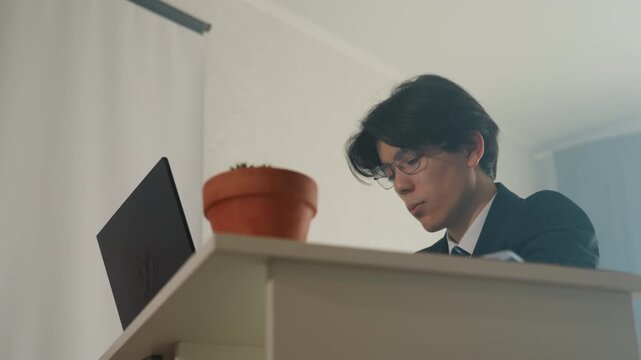 Asian analyst leaning over laptop in lowangle cinematic shot, potted plant on desk, wearing suit and glasses, reviewing spreadsheets and notes, focused expression, warm soft lighting, deadline