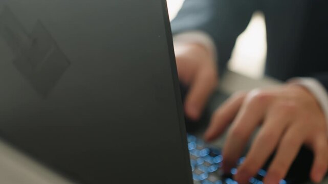 Closeup hands typing on laptop keyboard, suit sleeves visible, blue backlit keys glowing beneath fast keystrokes focused financial analyst composing emails and reports at office desk, modern