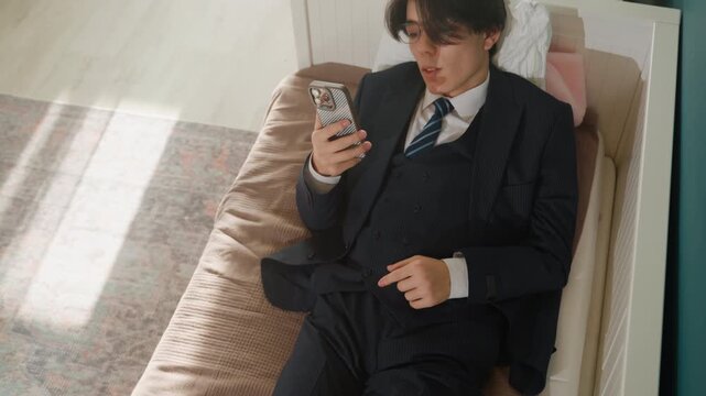 Asian businessman in suit on couch scrolling phone during quiet morning break, glasses pushed back, tie slightly loosened, sunlight casting soft shadows across carpet, shoes left beside couch, relaxed