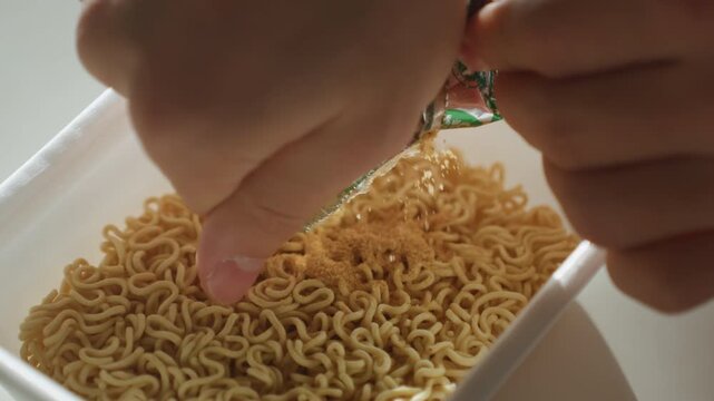 Hand sprinkling seasoning over dry ramen, closeup of instant noodles in plastic bowl as green foil packet is squeezed, seasoning flakes falling onto textured noodles, morning sunlight, cozy home