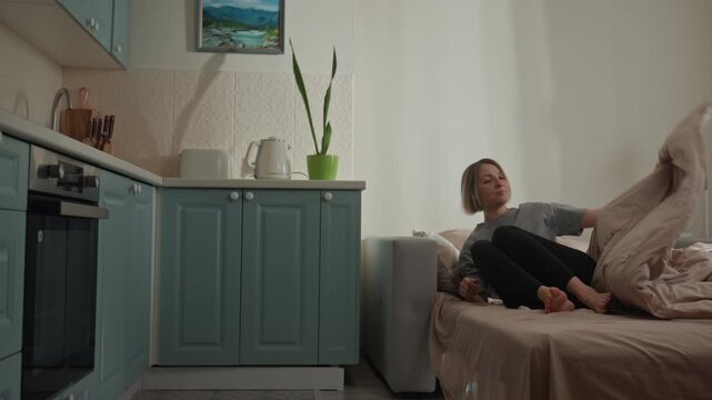 White woman wakes in studio bed soft morning light, pastel kitchen cabinets, green potted plant on counter music lover sits up, stretches, swings legs, stands and walks through compact apartment mood