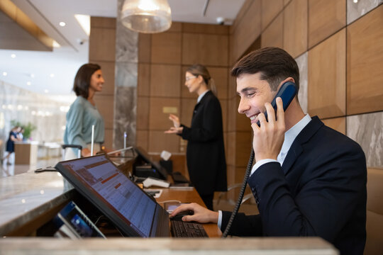Receptionist handling administrative tasks in reception front desk, talking to hotel guests on landline phone call, working with booking reservations. Customer services, inquiries management concept