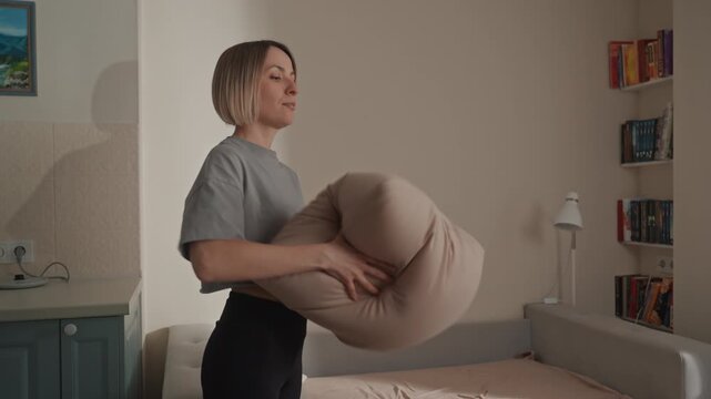 White woman holding pillow near bed, bookshelf backdrop, thoughtful expression, careful placement and gentle motions hinting music lover mood, casual grey shirt, soft neutral tones, indoor afternoon