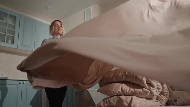 Young caucasian music lover tossing duvet with joyful rhythm in bright kitchenette bedroom, energetic movement while smoothing bedding, upbeat mood, candid smile, sunlight across pink linens, morning