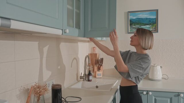 White woman in kitchen music lover taps speaker while reaching for upper cabinet and brewing coffee pastel cabinets, small black Bluetooth speaker on counter, ceramic mug, dried flowers, framed