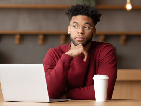  young man in glasses and a red sweater sits thoughtfully at a desk with a laptop and a takeaway coffee cup in a modern work environment.