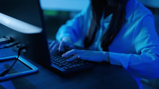Close up of anonymous female programmer typing on computer keyboard in dark office with blue neon light. Cybersecurity hacker working late at night on coding project. Data security and technology.