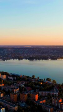 Vertical video. Russia, Votkinsk. Aerial drone panorama over the Votkinsk Reservoir embankment at sunset, warm golden light reflecting off calm water against a sprawling residential skyline. Drone fo