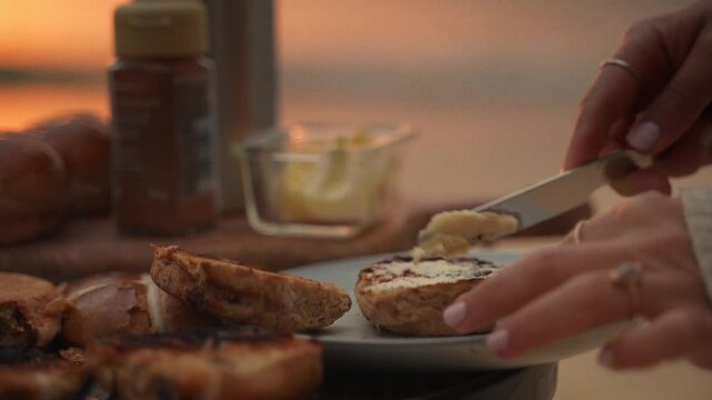 Morning Pastry Delight: A person spreads butter on a toasted bagel with a knife, preparing for a delightful breakfast against a soft-focus backdrop of a tranquil sunrise over a body of water.