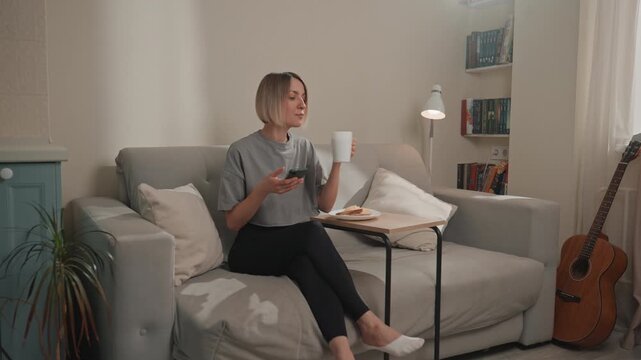 White woman scrolling phone with mug, enjoying breakfast pastry on side table, checking messages and tapping playlist, guitar leaning in corner, soft window light, playful smile, casual morning