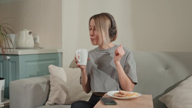 White woman with headphones sipping mug cozy living room morning ritual music lover enjoying coffee and toast, songwriter humming along, podcast host checking phone between bites, remote teacher