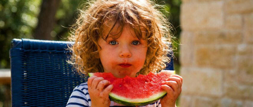 Cute toddler with curly hair eating a fresh slice of watermelon. Young child enjoying a healthy snack outdoors on a sunny day. Summer childhood concept, ideal for family nutrition marketing.