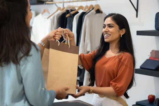 Young Indian female retail assistant gives paper shopping bag to woman client after successful purchase in modern clothing store. Customer services and satisfaction in shopping experience, consumerism