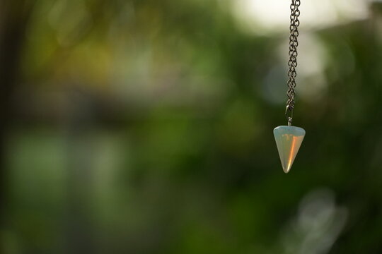 A conical opalite crystal pendulum hangs gently on a silver chain outdoors.