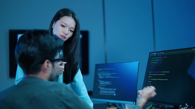 Two Asian software developers collaborating on code late at night. A male programmer points at the computer monitor while a female colleague analyzes data in a tech office. IT teamwork, coding.