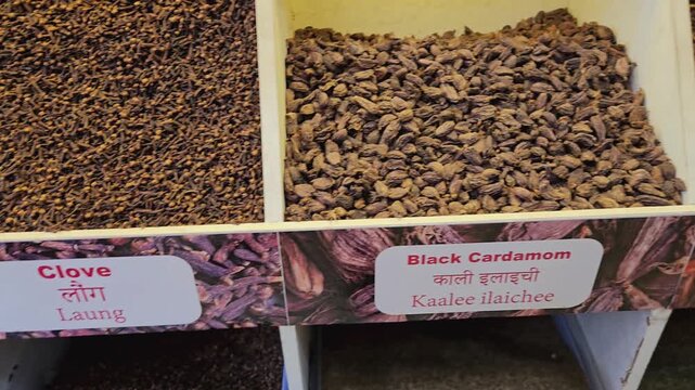 A vibrant display in a Kerala spice market features white pepper and cloves in large bins, complete with clear bilingual labels for shoppers looking for authentic regional ingredients