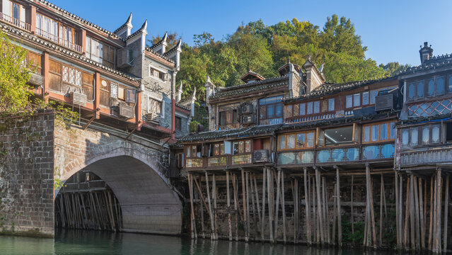 An ancient Chinese city Fenghuang. There are old wooden stilt houses on log piles along the shore. Arched stone bridge over the water. Green trees, clear blue sky. China. Phoenix. Tuojiang River