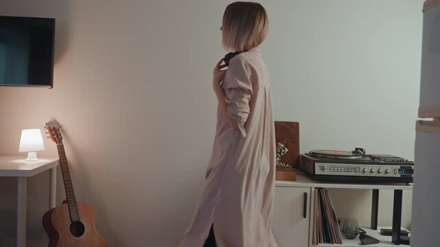 Blonde woman picks record in intimate setting. Young lady with guitar selects vinyl in warm studio corner. Female vinyl enthusiast in comfortable studio space with guitar and soft lighting