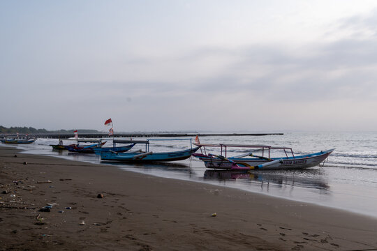 Cilacap, Indonesia - May 13, 2026: Colorful traditional outrigger fishing boats lined up along the shore of Teluk Penyu Beach under an overcast sky.
