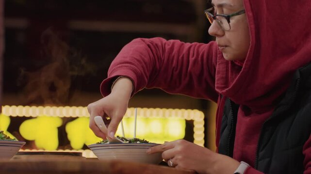 Night neon counter steaming bowl ramen savoring soup, hooded diner under marquee lights adding spoon and chopsticks, closeup profile with glasses, warm broth steam rising, lively market backdrop,