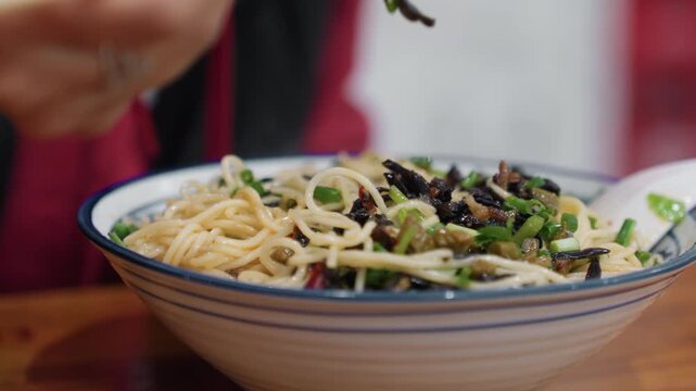 Closeup bowl noodles chopsticks hands lifting ramen strands, scallion and seaweed garnish on warm wooden table, savory broth glistening in ceramic bowl, slow intimate eating motion, casual eatery