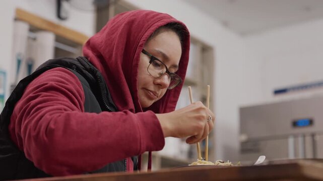 Female sampling noodles with chopsticks in test kitchen, red hoodie and glasses, leaning over counter, focused expression, tasting for texture and flavor, industrial shelves and utensils