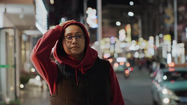 Night street portrait woman student hoodie glasses, contemplative expression amid neon signs and blurred traffic, standing on busy sidewalk, reflective mood, soft bokeh, downtown lights, casual red