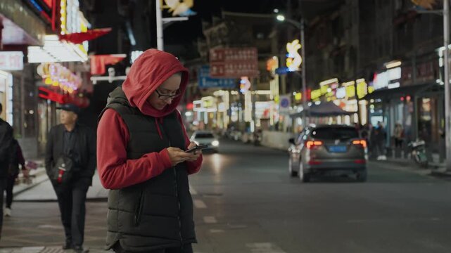 Asian woman walking at night in neon street checking phone, wearing red hoodie and black vest, passing cars and illuminated storefront signs, solitary student pausing to read message, cinematic bokeh