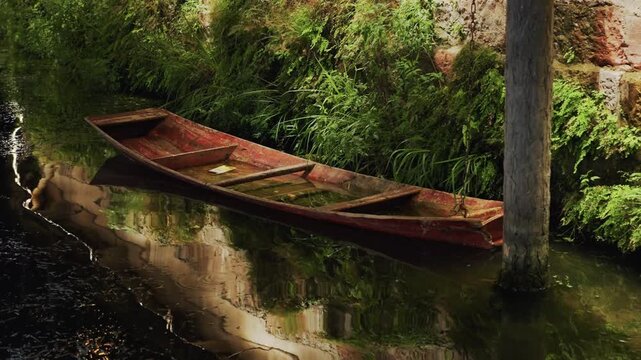 Night river walk, abandoned wooden skiff moored beside mossy brick wall, still water reflecting dense foliage, leaning post and dark ripples, atmospheric quiet and nostalgic decay in gentle twilight.