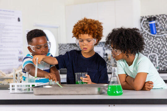 Diverse children wearing goggles mixing green solution with beaker and tubes at lab bench