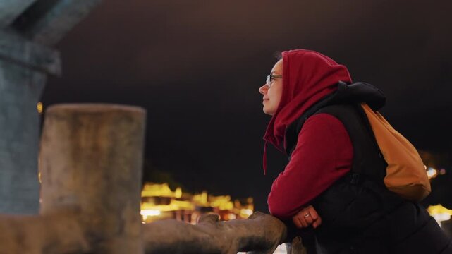 Night riverfront skyline woman observing from stony quay with hands on bollard, distant bridge lights and lantern glow, orange backpack and red scarf, quiet travelers pause, candid urban evening