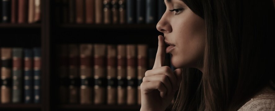 Woman puts finger to lips in library silence. Woman makes shushing gesture near bookshelves, asking for quiet. Discreet communication, secret knowledge, private conversation. Person asks for hush.