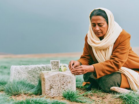South Asian woman kneeling at an outdoor graveside tribute placing a flower and seed card for bereavement support, eco memorial services and grief counseling editorial
