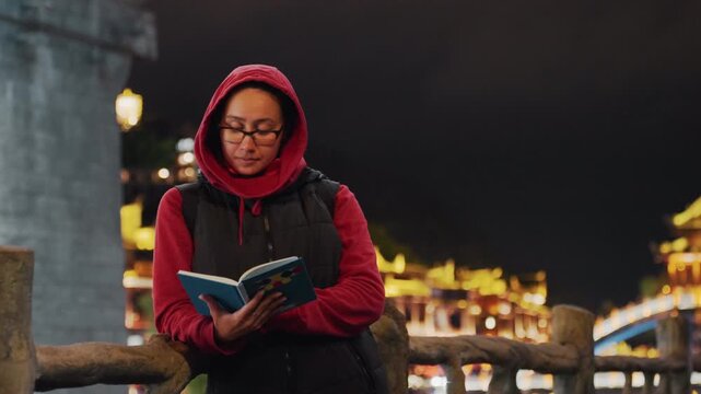 Asian woman reading book on bridge at night, urban explorer vibe peering at pages between strolls, waterfront lantern reflections and illuminated arches, travel curiosity under city lights