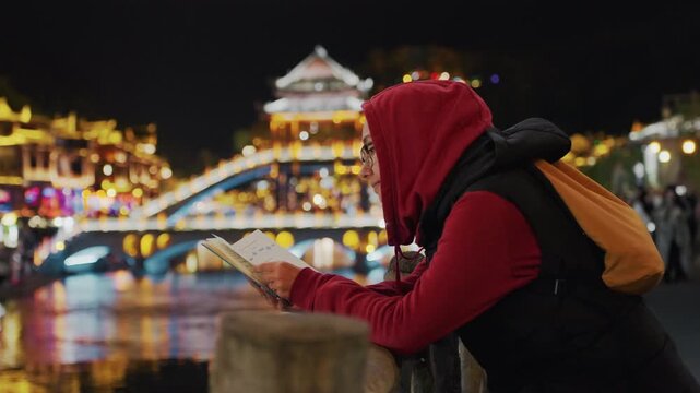 Night bridge lights reflecting over river, student in red hoodie leans on railing reading map under warm bokeh peaceful waterfront stroll scene, soft glow from lanterns, distant architecture and calm