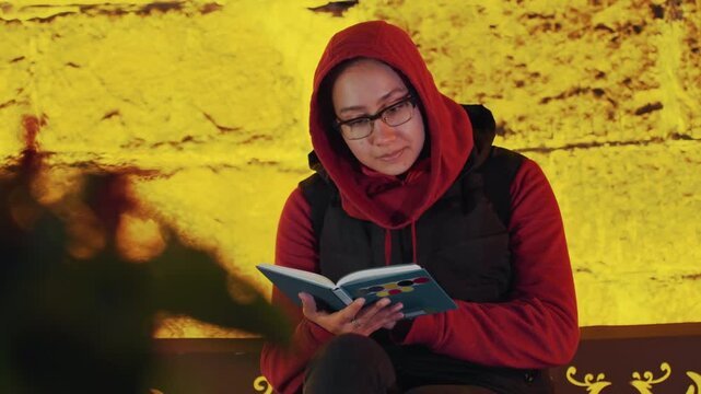 Quiet woman savoring book moment. Relaxed woman with glasses enjoys tranquil evening with her book. Serene woman quietly indulges in her favorite book adventure near softly glowing wall